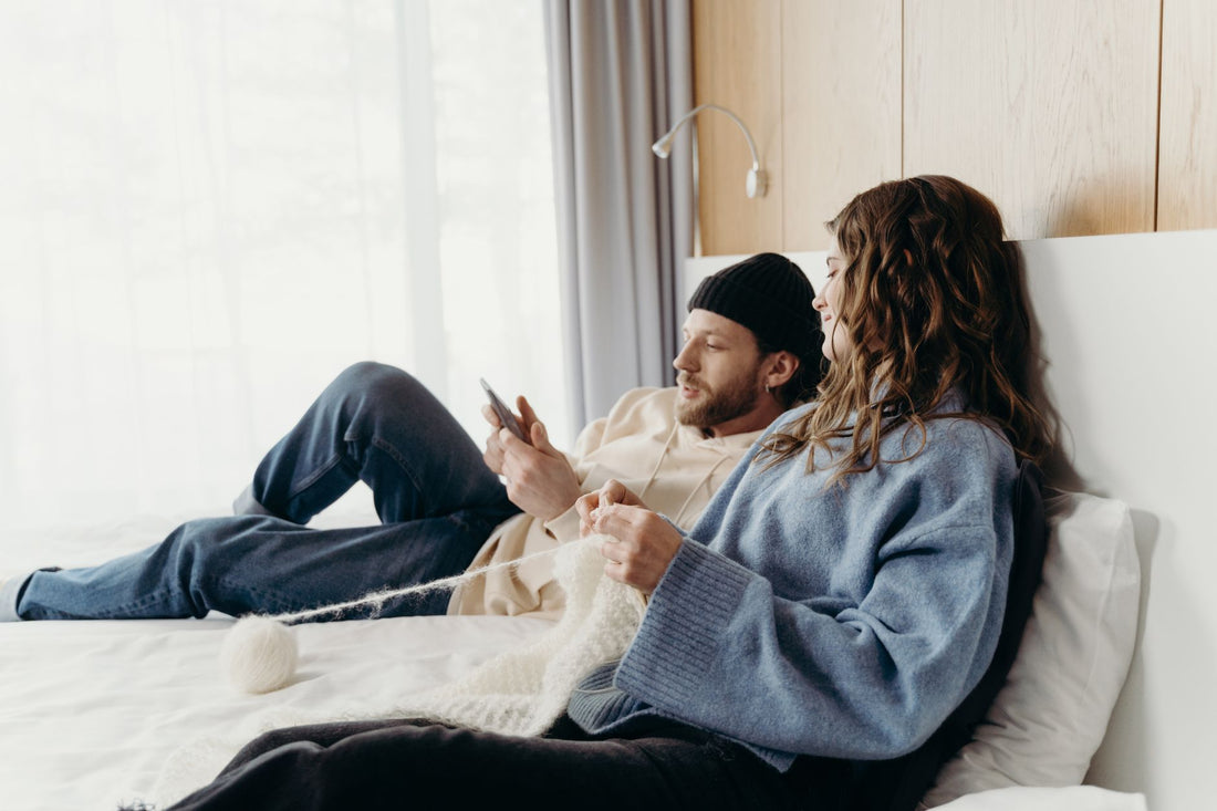 A couple relaxing in a cozy bedroom; woman knitting and man using smartphone.