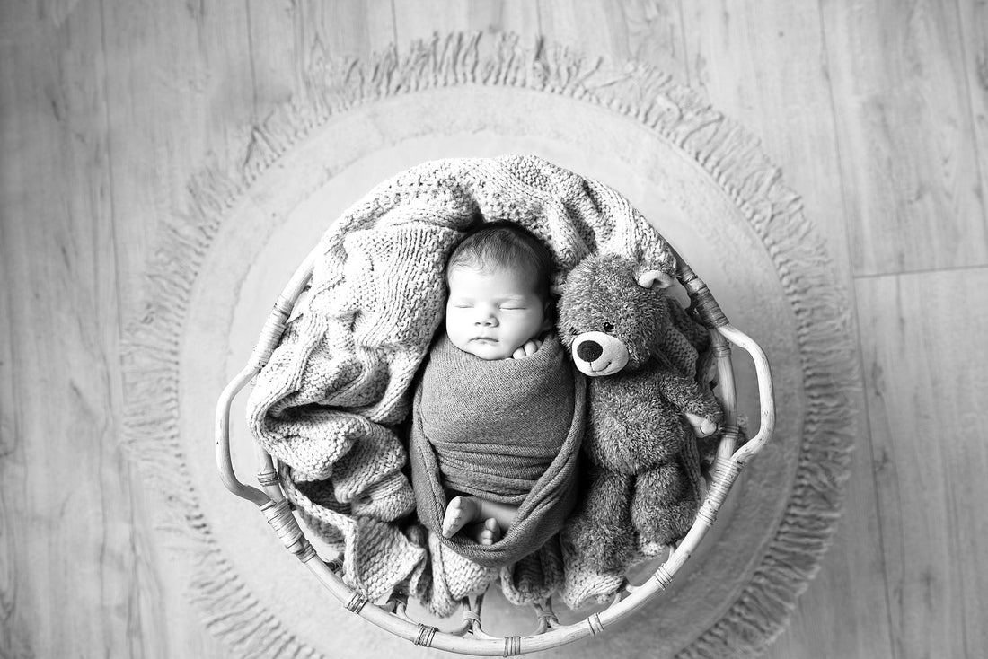 Black and white image of a baby swaddled in a basket with a teddy bear, exuding warmth and serenity.
