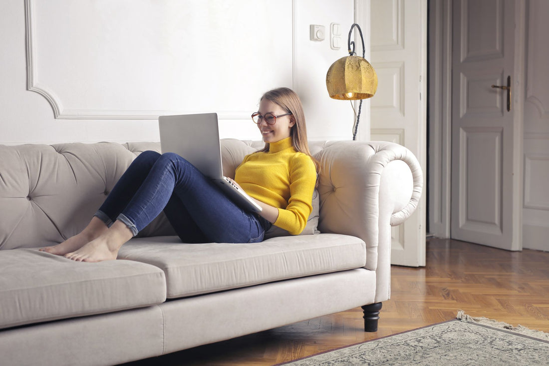 A woman in a yellow sweater uses her laptop while relaxing on a sofa at home.