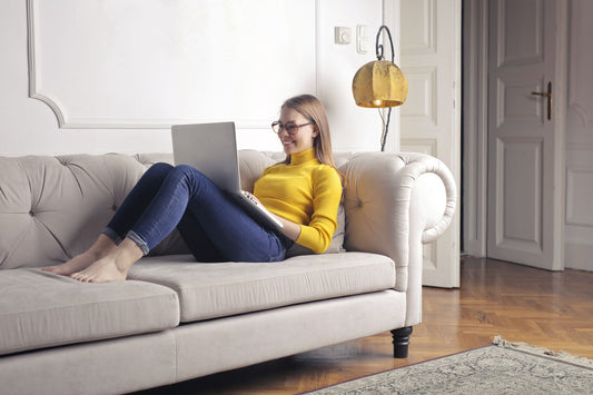 A woman in a yellow sweater uses her laptop while relaxing on a sofa at home.