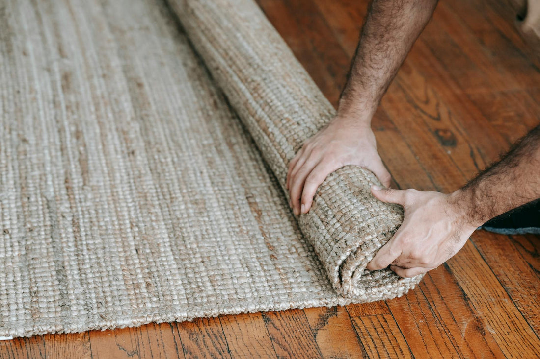 A man rolls up a textured woven rug on a wooden floor, indicating moving or cleaning.