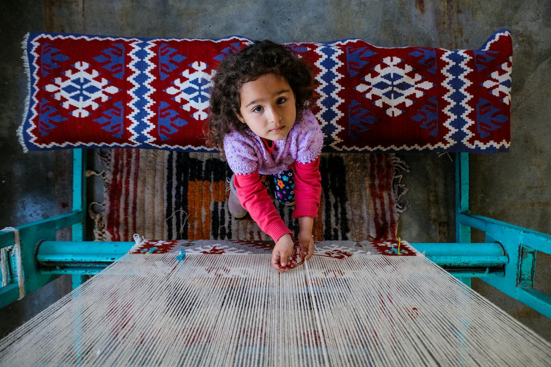 A young girl in colorful attire weaving a patterned rug on a traditional loom.
