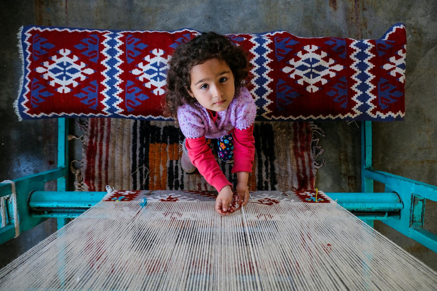 A young girl in colorful attire weaving a patterned rug on a traditional loom.