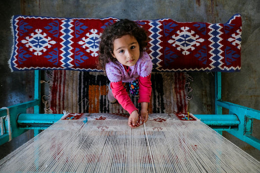 A young girl in colorful attire weaving a patterned rug on a traditional loom.