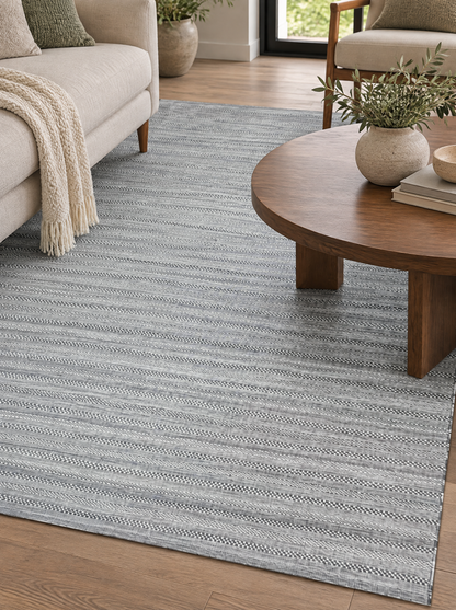 Artisan Eco Weave Grey Style Shot — Close view of a textured grey area rug beside a cream sofa and round wooden coffee table.